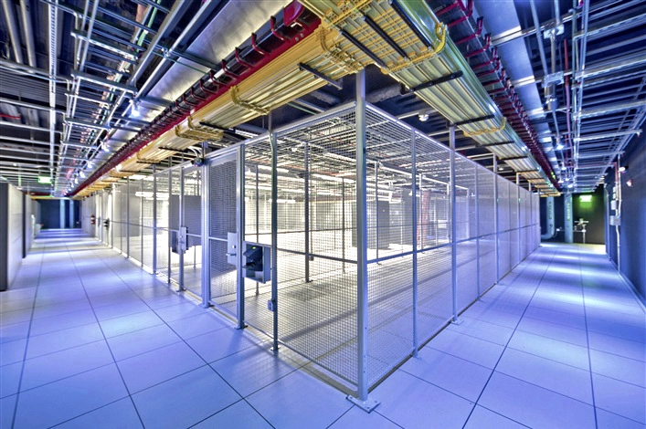Interior view of an empty data center server room hall, showing secure wire mesh cages, a white raised floor tile system, and overhead cable trays filled with multi-colored networking wires.