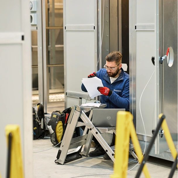 A Munters technician reviewing paperwork next to a laptop computer while standing on a small step ladder inside a manufacturing facility.
