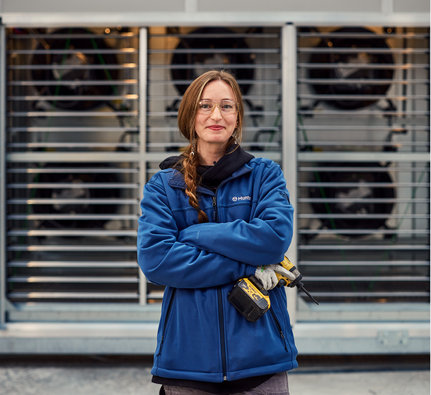 A smiling female engineer wearing safety glasses and a blue Munters winter jacket holding a cordless power drill, standing proudly in front of a Munters Fan Wall Unit (FWU) assembly.